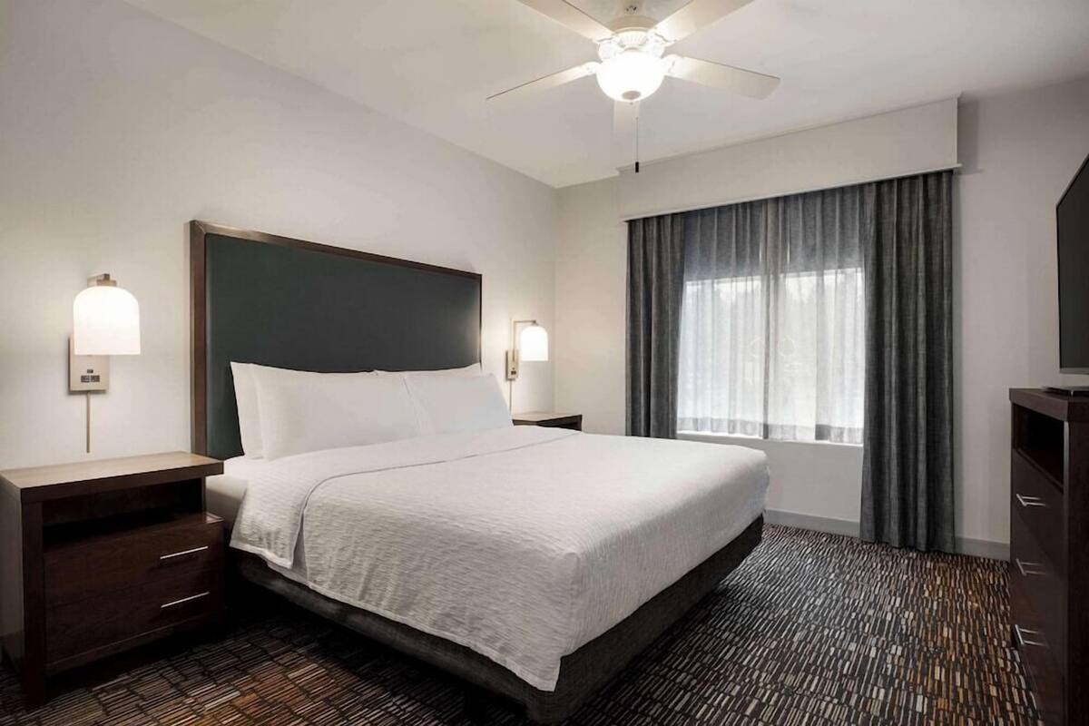 A hotel room with a large bed, two bedside tables, a ceiling fan, and a window with curtains.