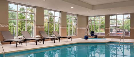Una piscina con sillas de descanso y una escalera, ventanas amplias y vistas a árboles y un estacionamiento.
