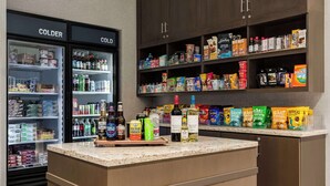 A kitchenette with a refrigerator, shelves stocked with snacks, and a countertop with various beverages and food items.