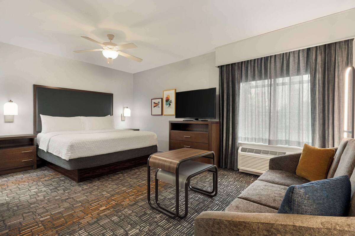 A hotel room with a bed, a sofa, a desk, a TV, and a ceiling fan.