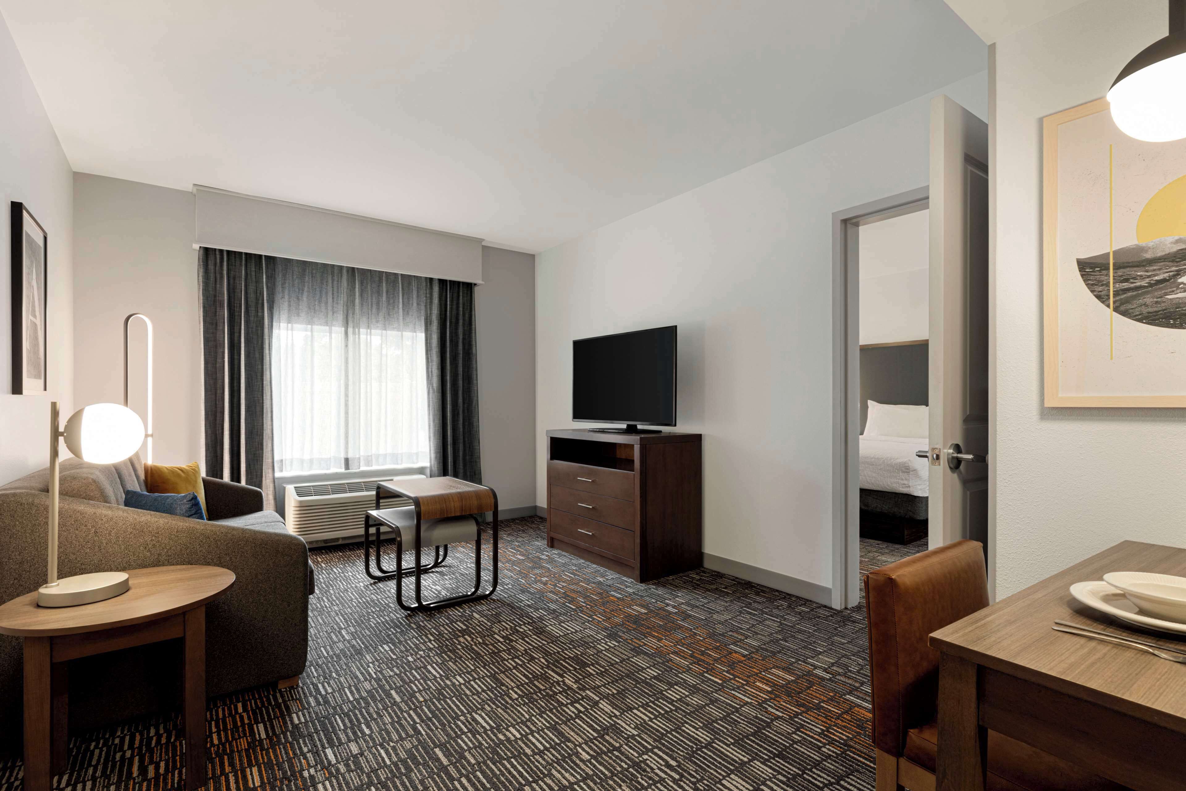 A hotel room with a sofa, a small table, a TV on a wooden cabinet, a dining table with chairs, and a large window with curtains.