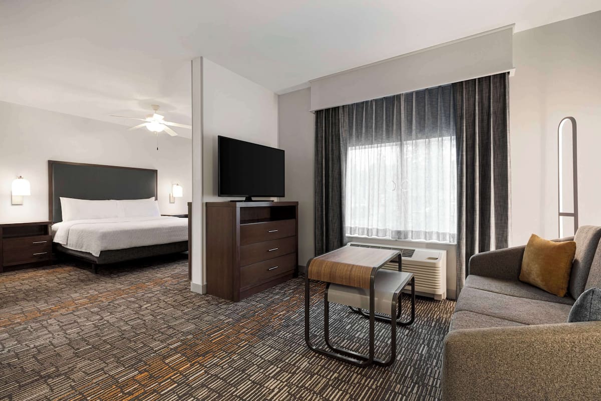 A hotel room with a bed, a TV, a desk, a sofa, and a large window with curtains.