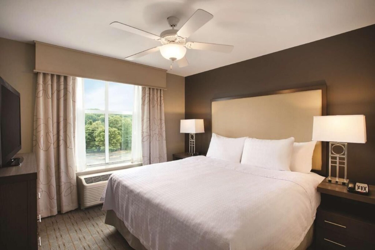 A hotel room with a large bed, two bedside lamps, a ceiling fan, a TV, and a window with curtains.