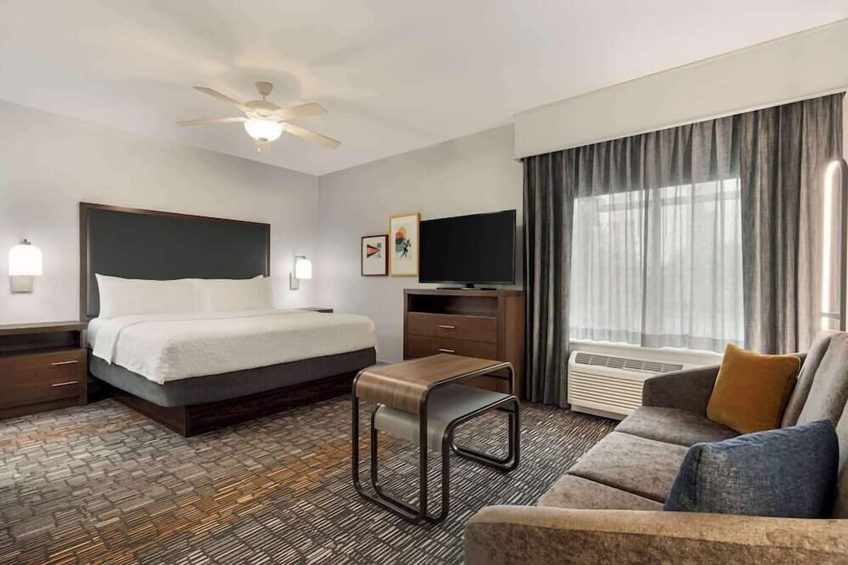 A hotel room with a bed, a sofa, a TV, a ceiling fan, and a window with curtains.