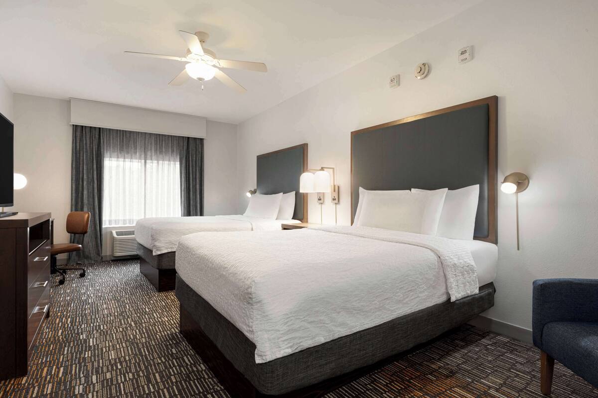 A hotel room with two beds, a desk, a chair, a ceiling fan, and a large window with curtains.
