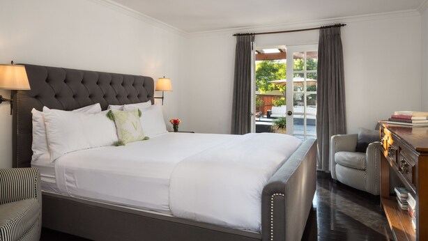 A neatly arranged hotel room with a large bed, a bedside table with a lamp, a striped armchair, and a view of the outdoors through a door. - The Charlie Hotel (West Hollywood)