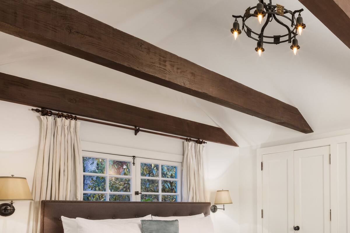 A bedroom with a large bed, wooden beams, a chandelier, a window with curtains, and a door.