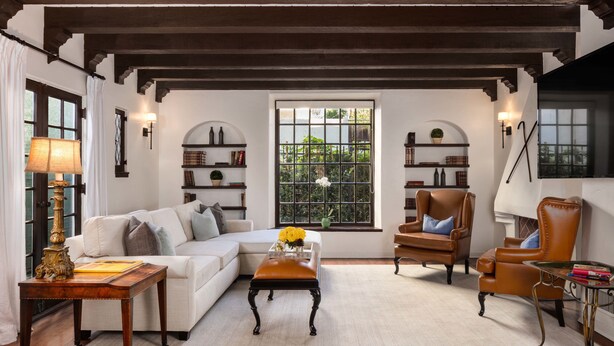 A cozy living room with a white sofa, a brown leather armchair, and a wooden coffee table. There are bookshelves with books and decorative items, and a window with a view of greenery. - The Charlie Hotel (West Hollywood)