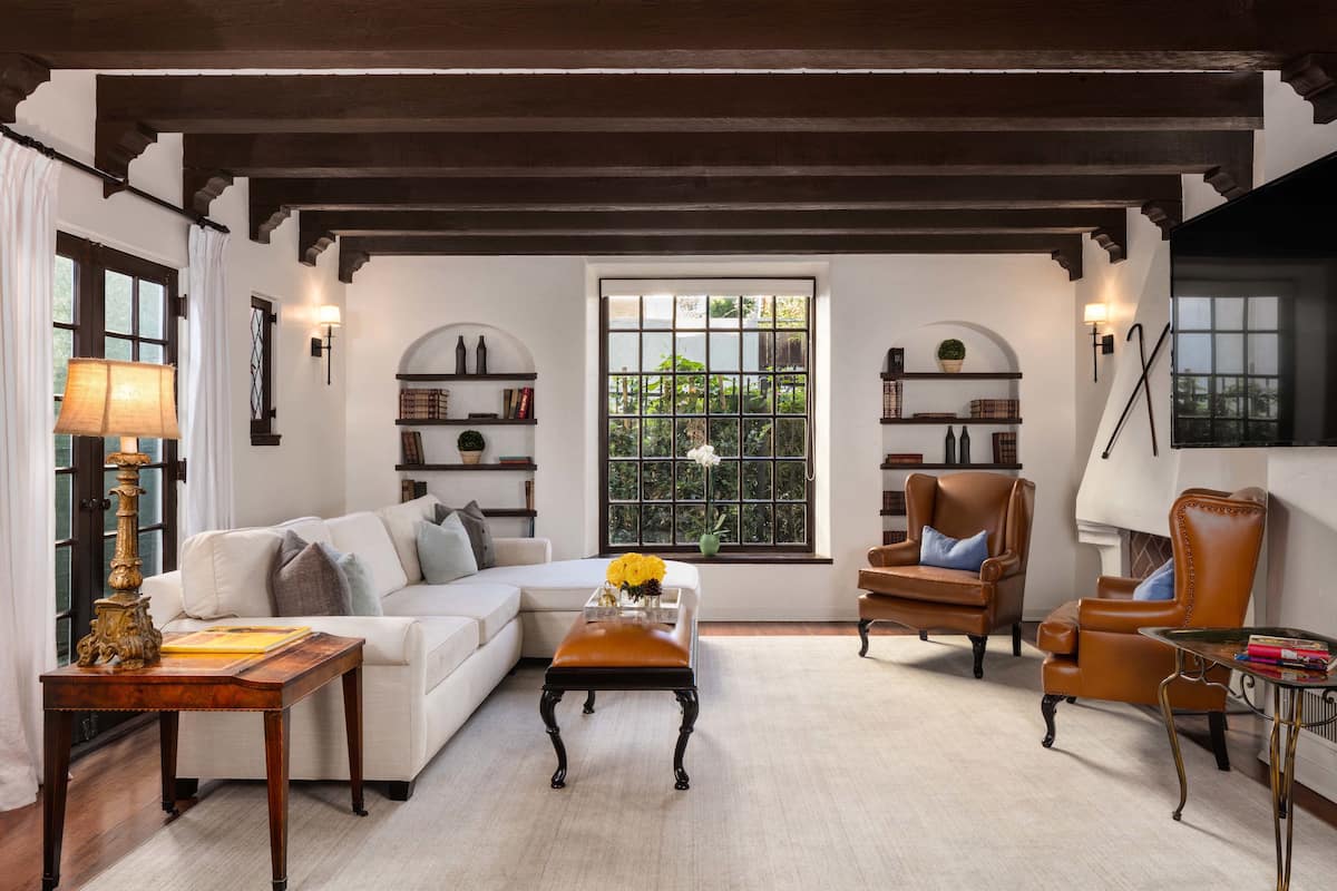 A cozy living room with a white sofa, a brown leather armchair, and a wooden coffee table. There are bookshelves with books and decorative items, and a window with a view of greenery.