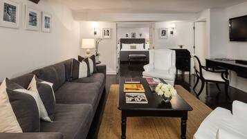 Een moderne woonkamer met een donkergrijze bank, een zwarte salontafel met boeken en bloemen, een witte fauteuil, en een bed op de achtergrond.