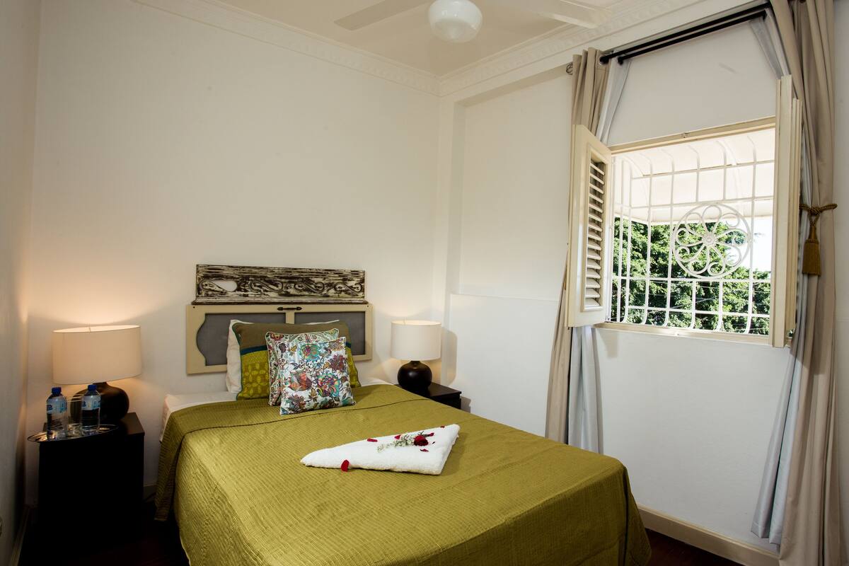 A neatly made bed with a green bedspread, two bedside lamps, a floral pillow, and a window with shutters.