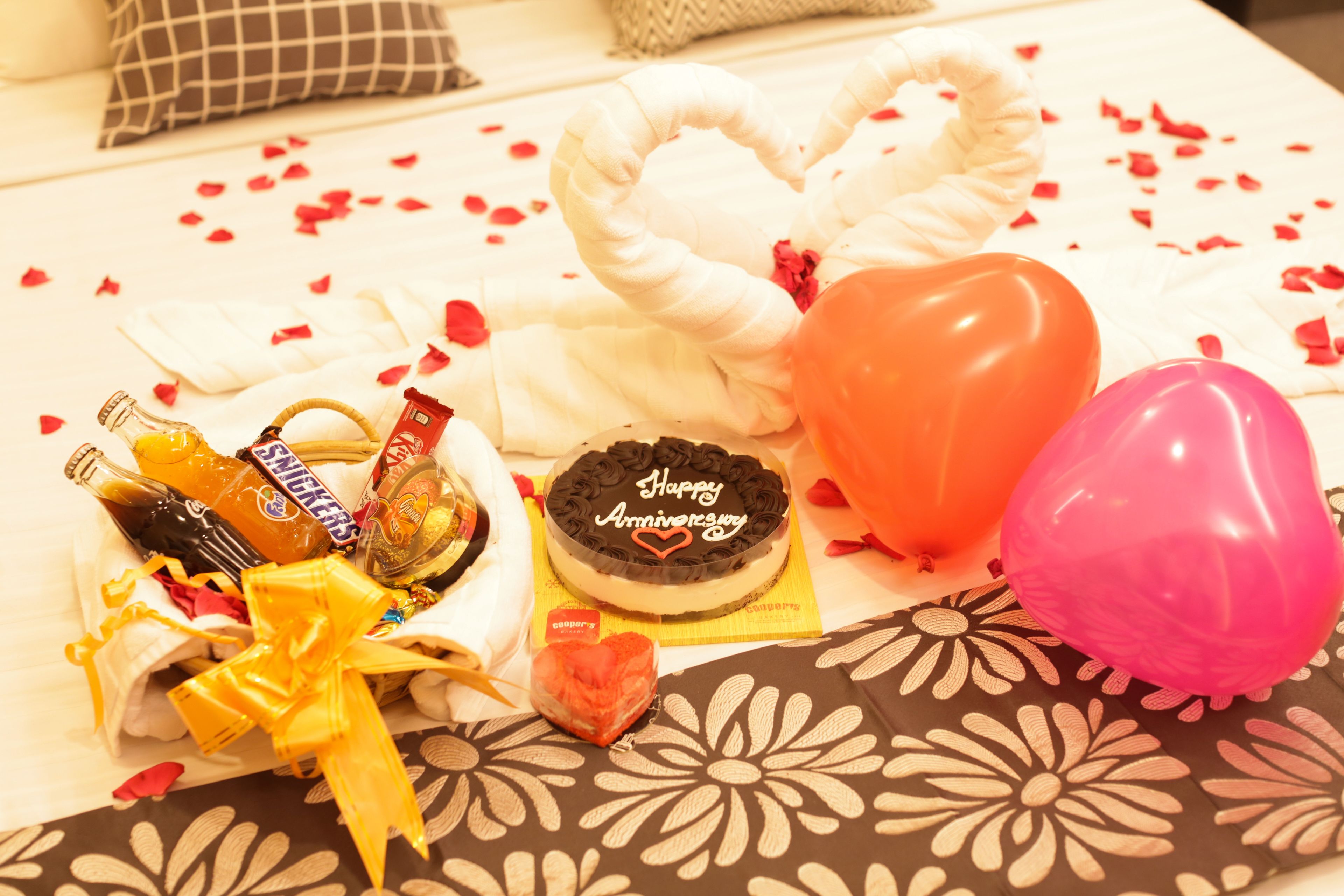 Una cama con un arreglo de almohadillas blancas con forma de corazón, un pastel de chocolate con la inscripción 'Feliz Aniversario', un ramo de flores y un globo rosa.