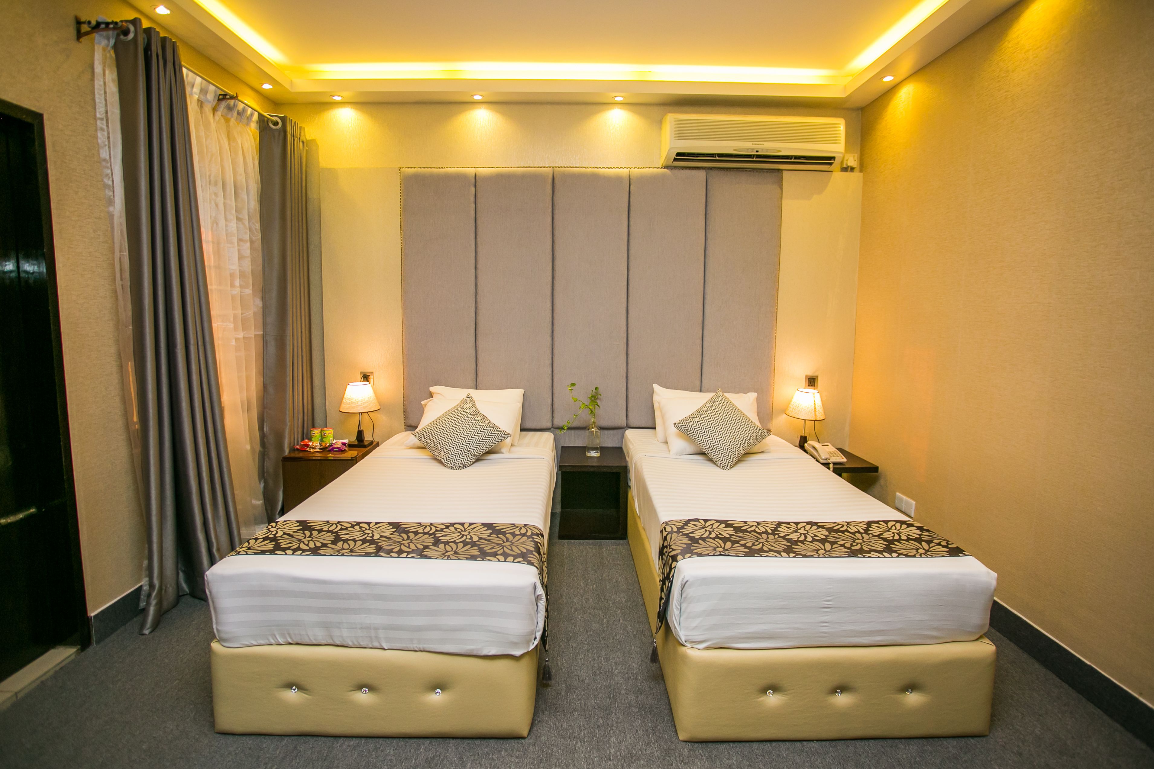 A hotel room with two beds, a large window, an air conditioning unit, and a nightstand with a lamp.
