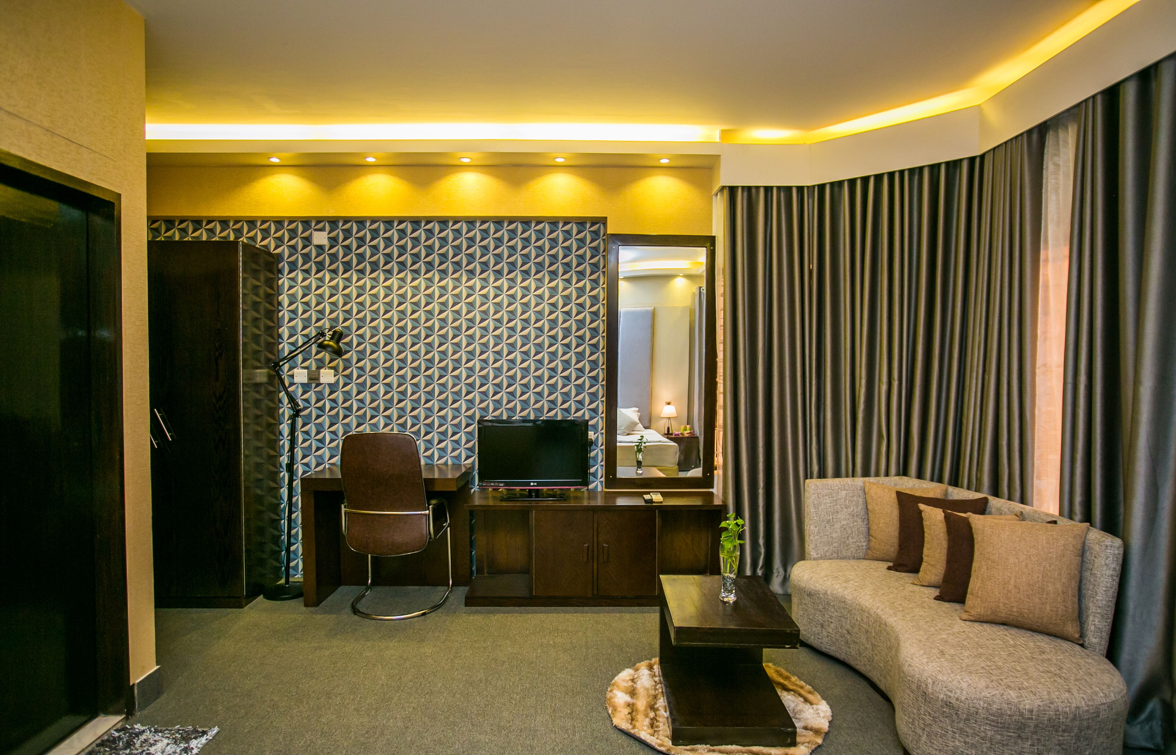 A modern hotel room with a sofa, a TV, a desk, and a patterned wall.