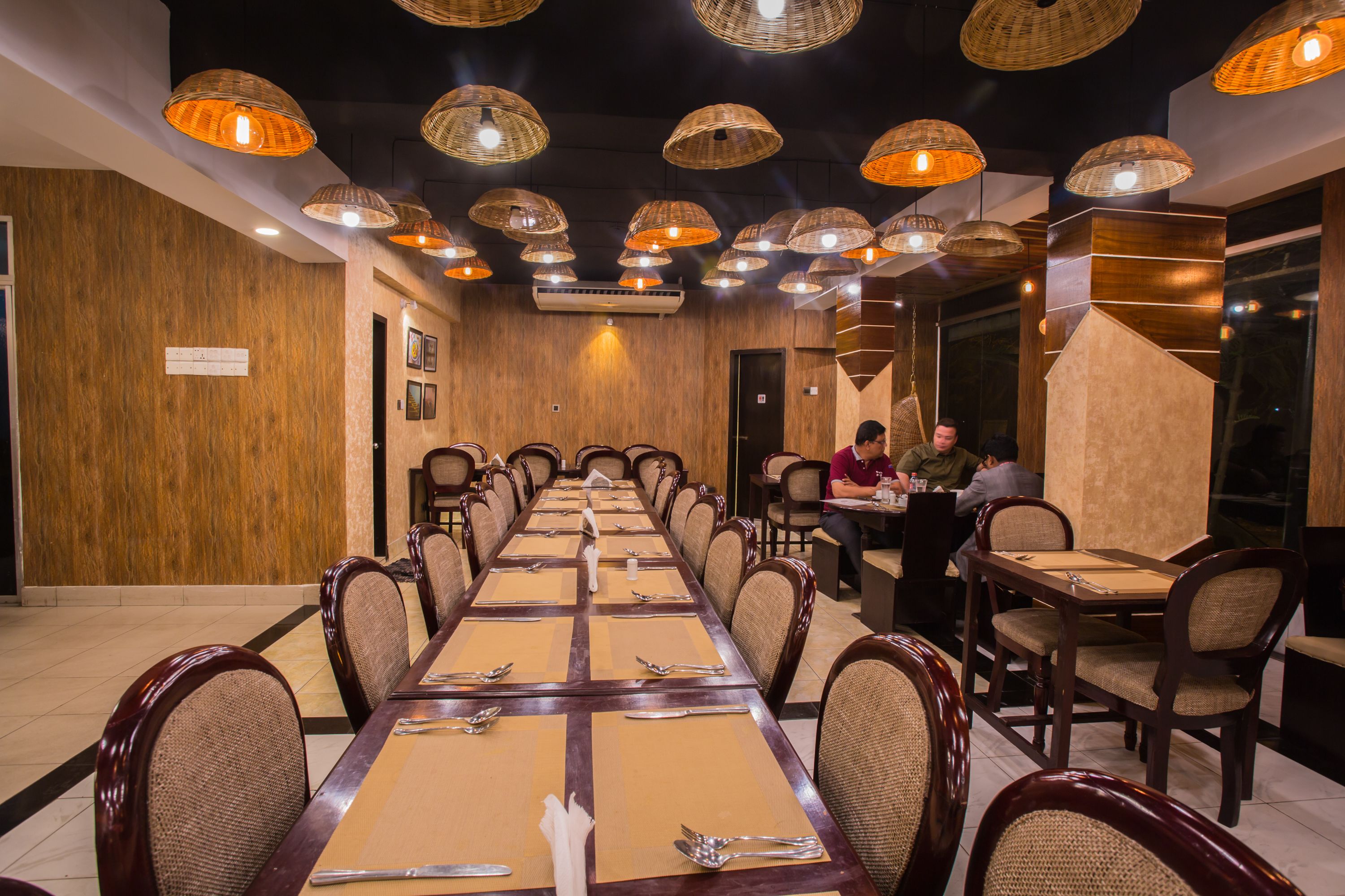 A long dining table with multiple chairs, overhead wicker lamps, and a wooden wall.