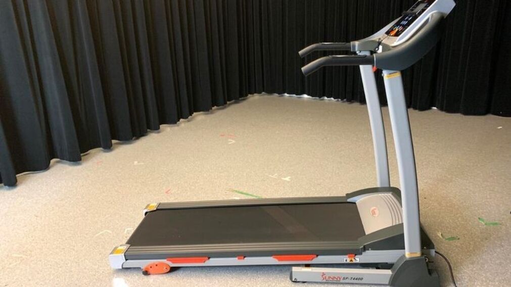 A treadmill with a control panel, placed on a carpeted floor with a black curtain in the background.