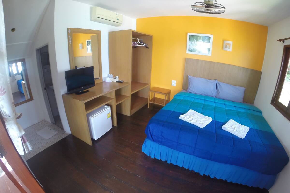 A bedroom with a bed, a desk, a chair, a wardrobe, a television, and an air conditioning unit.