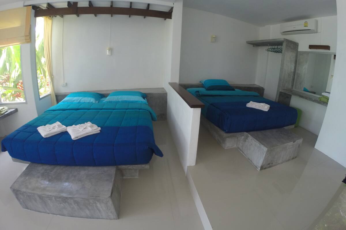 A room with two beds, each with a blue bedspread and pillows, and a small table with a mirror and a shelf.