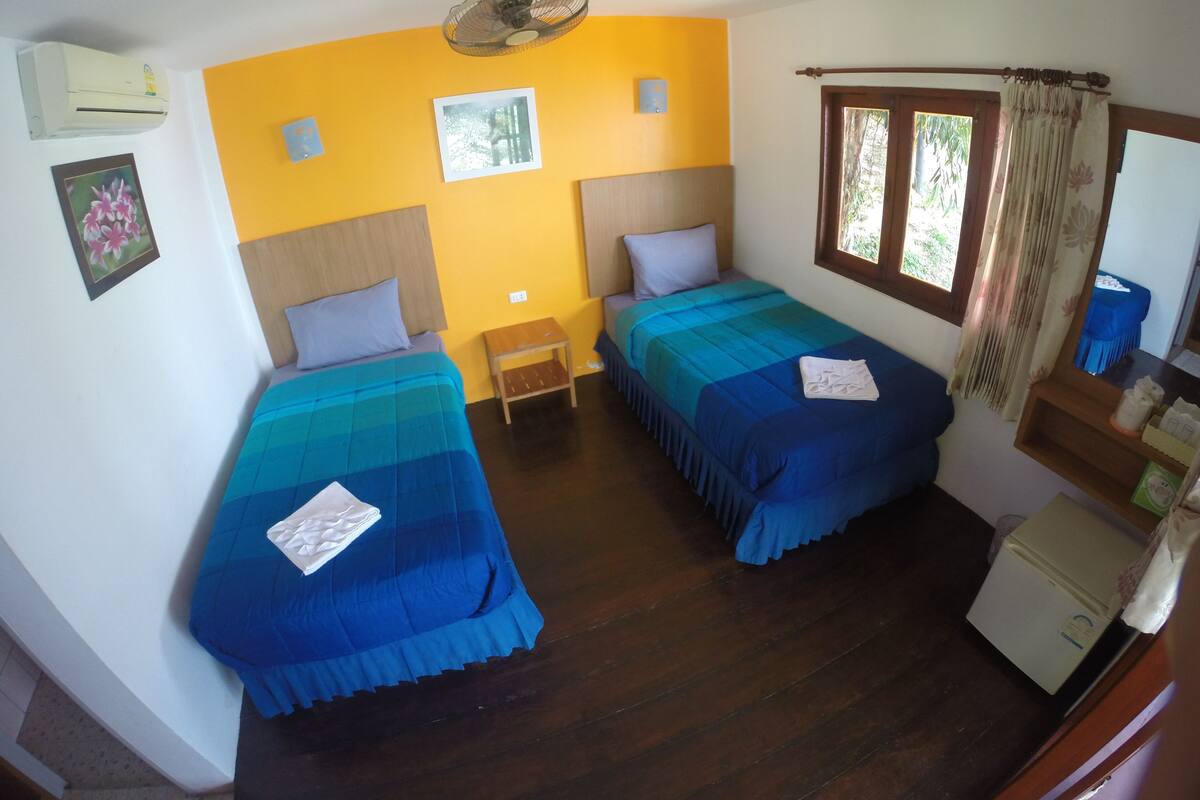 A room with two beds, a small table, a ceiling fan, and a window with curtains.