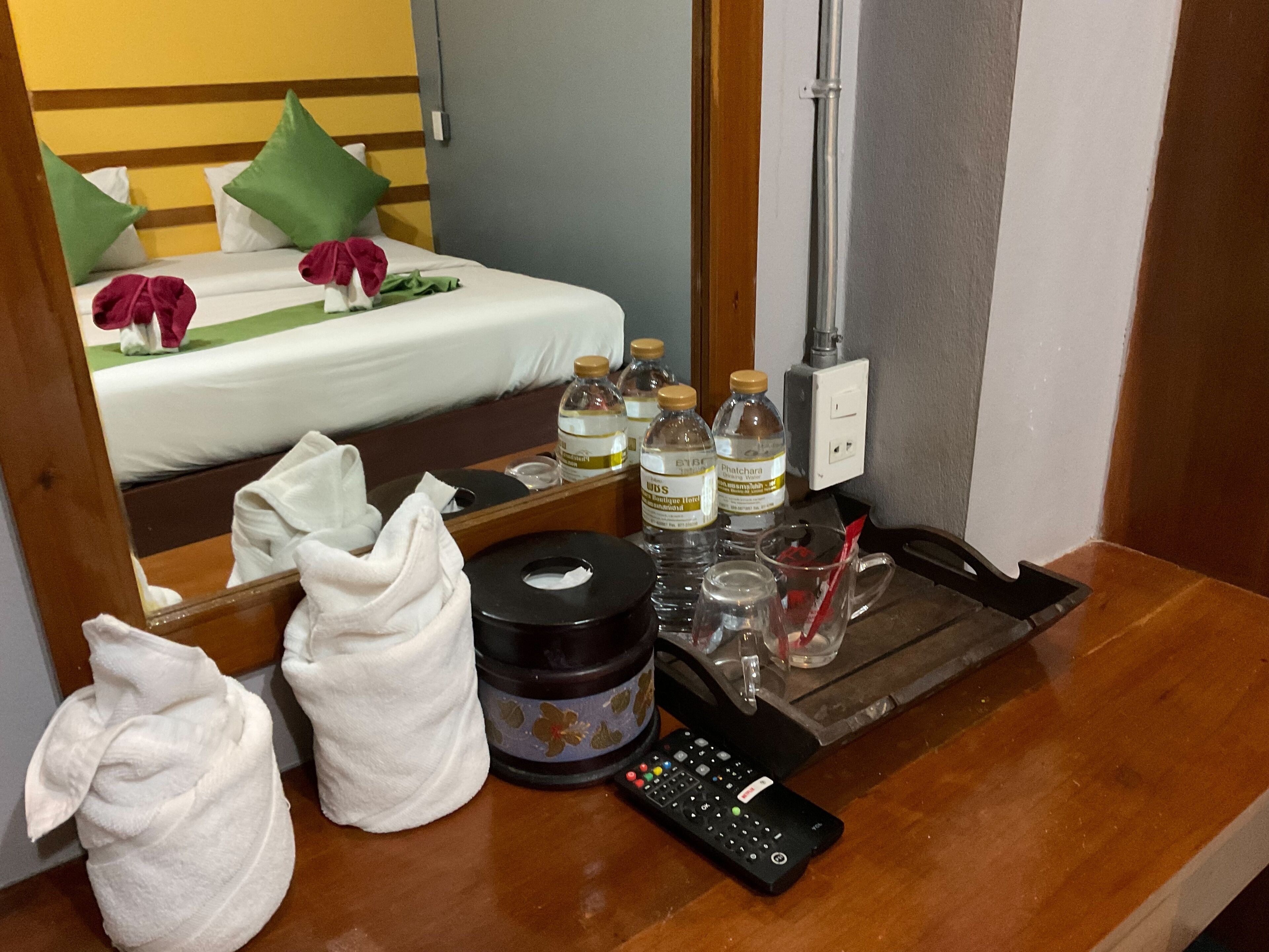 A hotel room with a bed, a mirror, a wooden side table with toiletries, a remote control, and a minibar.
