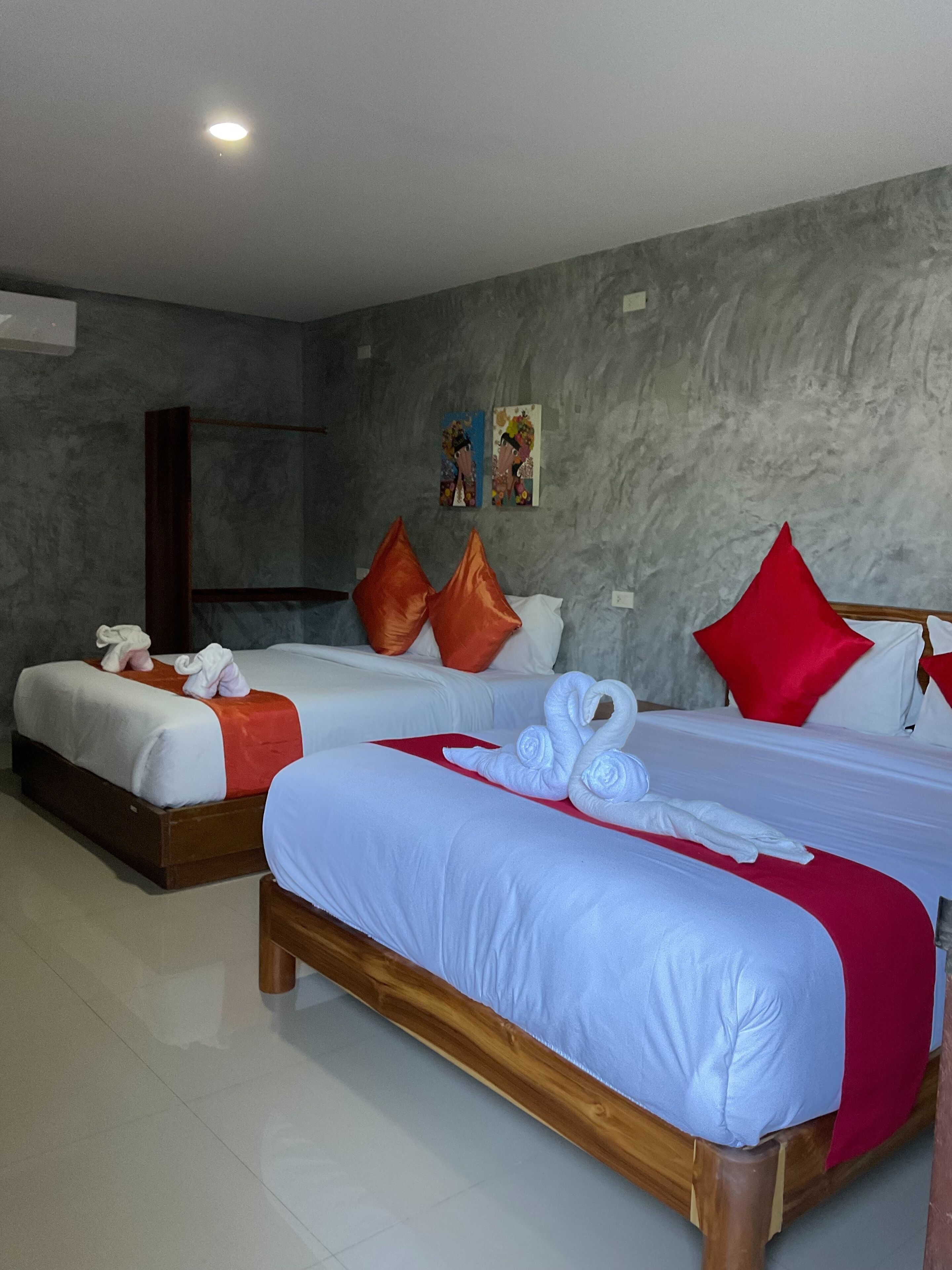 A hotel room with two beds, each with white linens and red accents, a grey textured wall, and a wooden floor.