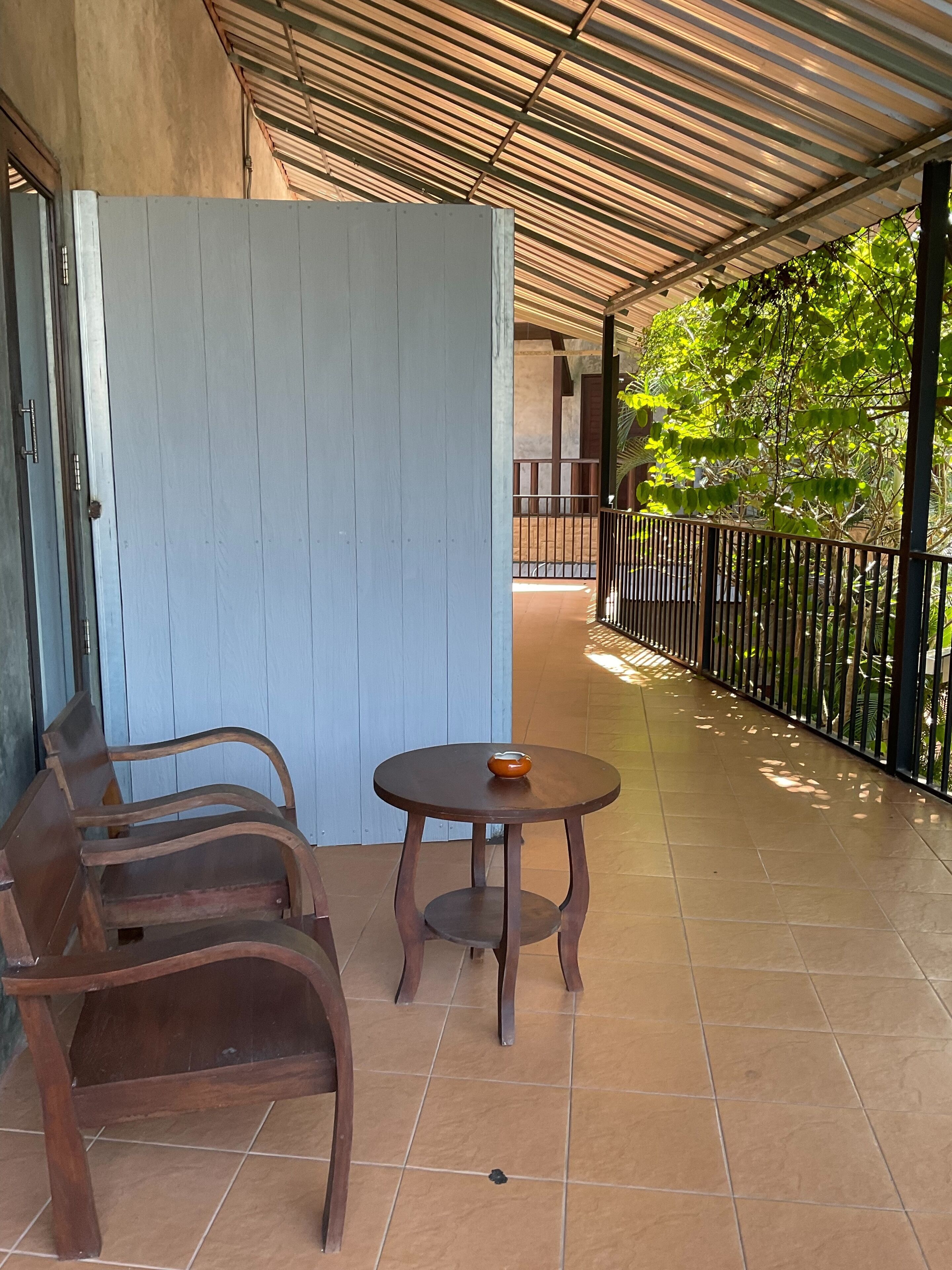 Uma área de estar coberta ao ar livre, com um banco de madeira e uma pequena mesa, piso de azulejo e corrimão de metal.