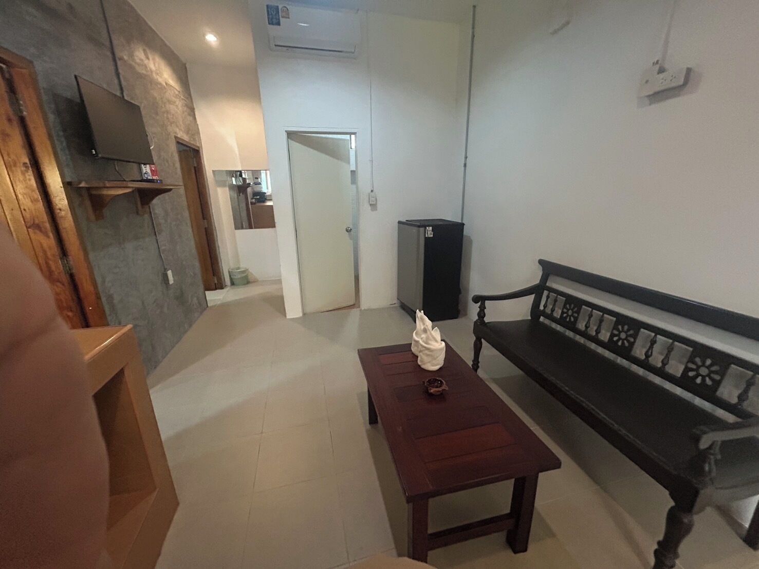 A living room with a wooden bench, a small table with a decorative item, a television on the wall, and a door leading to another room.