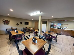 A dining area with multiple tables and chairs, a kitchen area with a refrigerator and coffee machine, and a wall-mounted television.