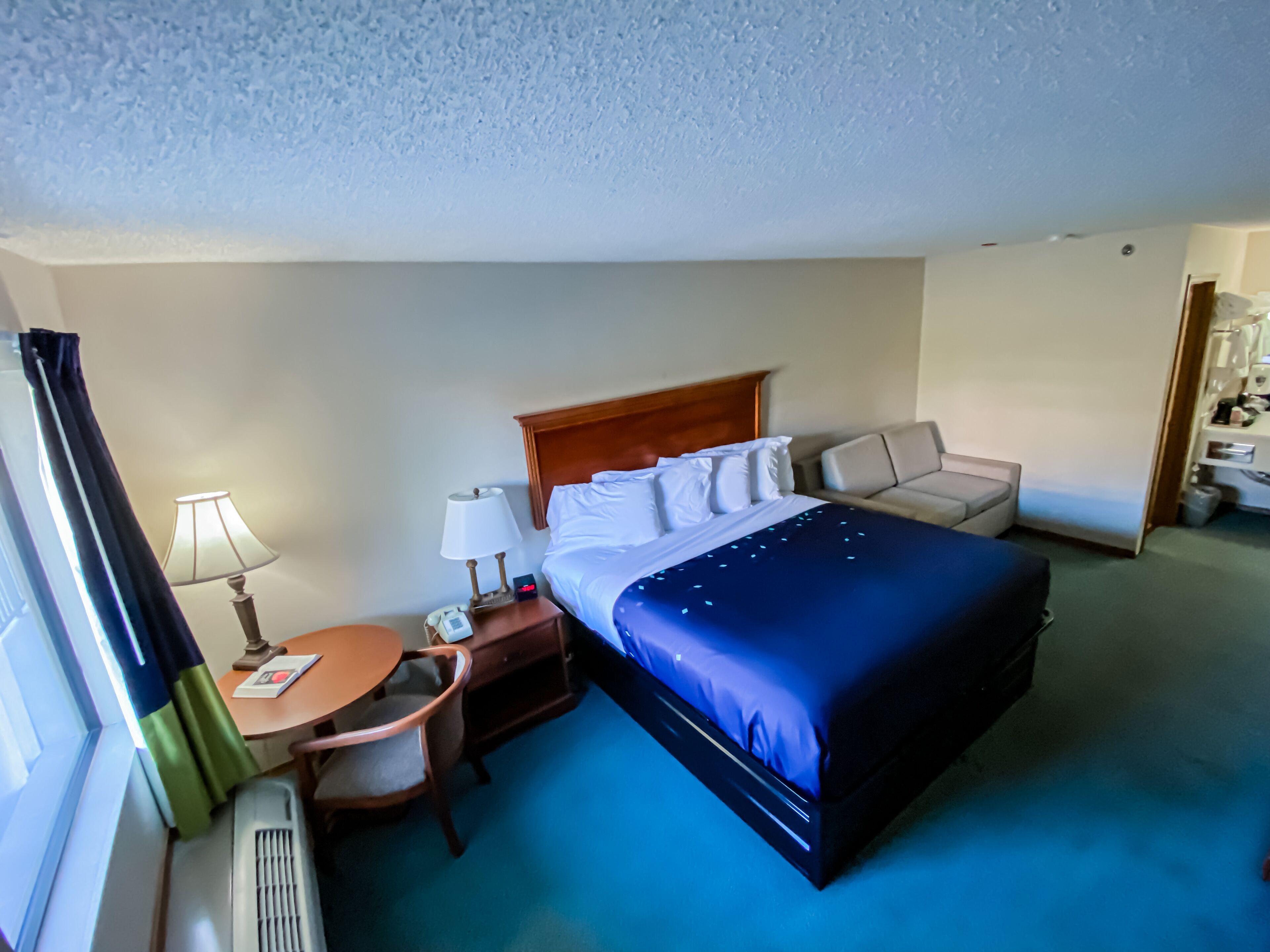 Habitación de hotel con cama, escritorio, silla, lámpara, mesita de noche y ventana con cortinas.