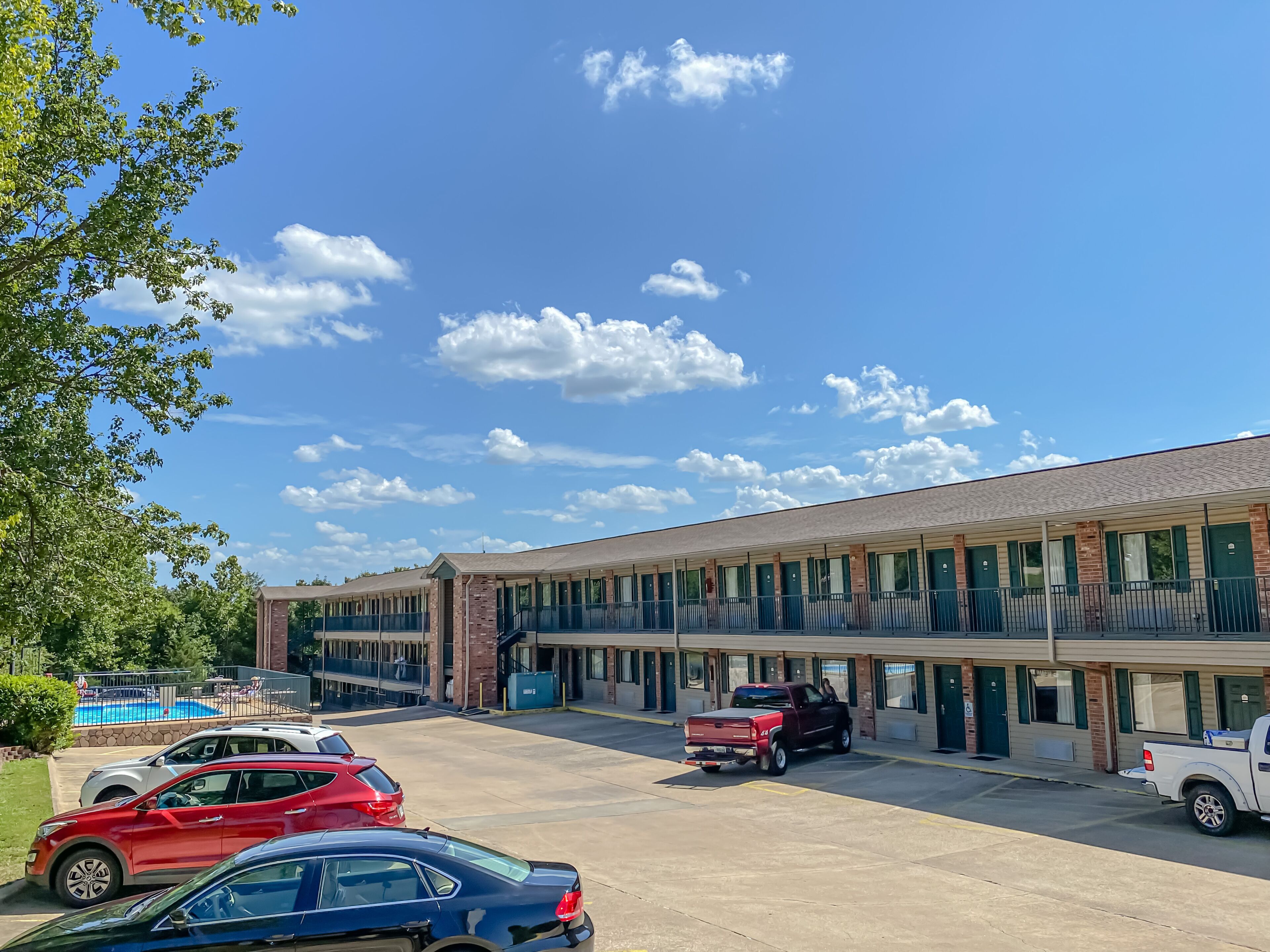 Un hôtel doté d’un parking, où sont garées plusieurs voitures, et un ciel bleu azur parsemé de nuages.