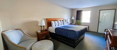 Quarto de hotel com uma cama, uma cadeira, uma escrivaninha e um pequeno banquinho.