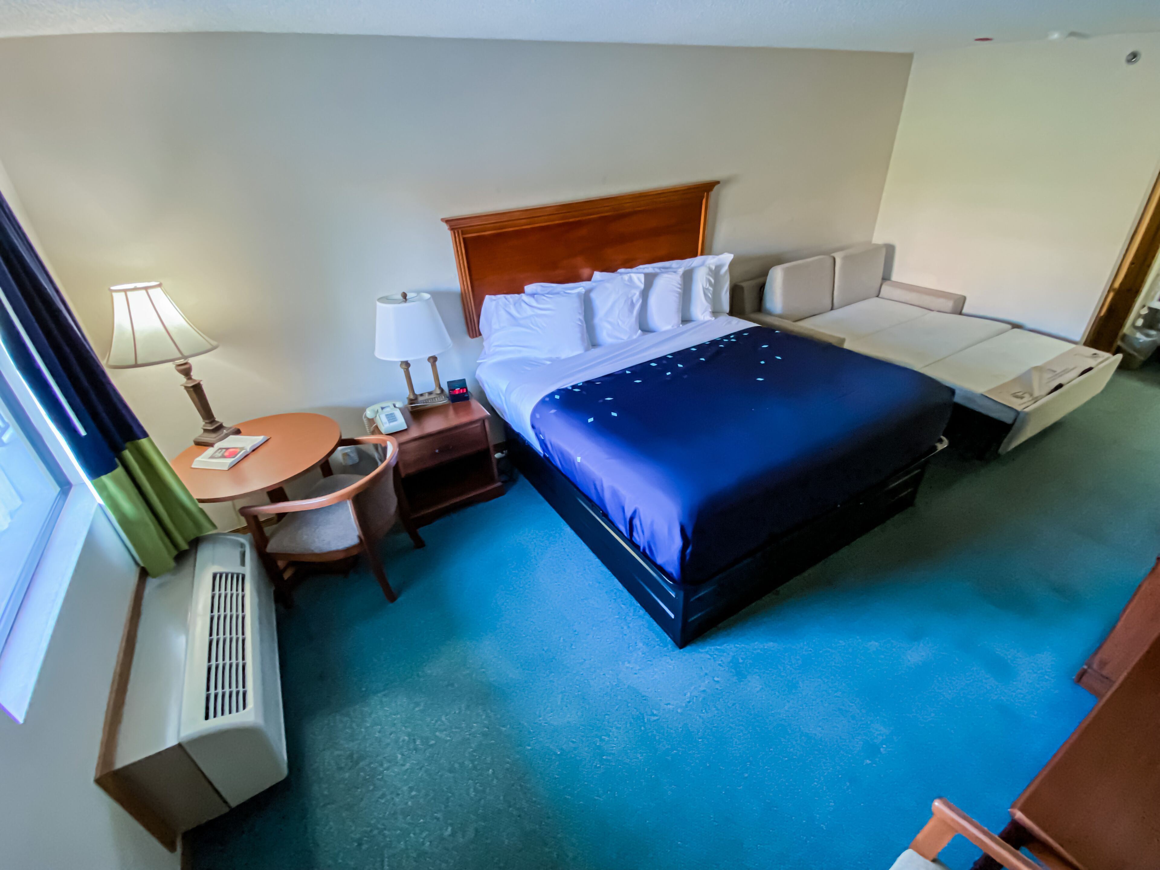 A hotel room with a large bed, a bedside table with a lamp, a small round table, and a chair.