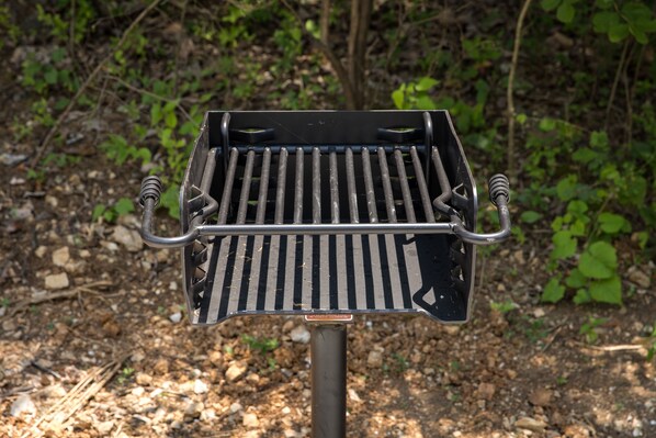 A barbecue grill with a metal grate and two handles, set on a stand in a wooded area.