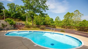 A swimming pool with a curved edge, surrounded by a metal fence, and set against a backdrop of trees and a clear sky. - Rosebud Inn (Branson)