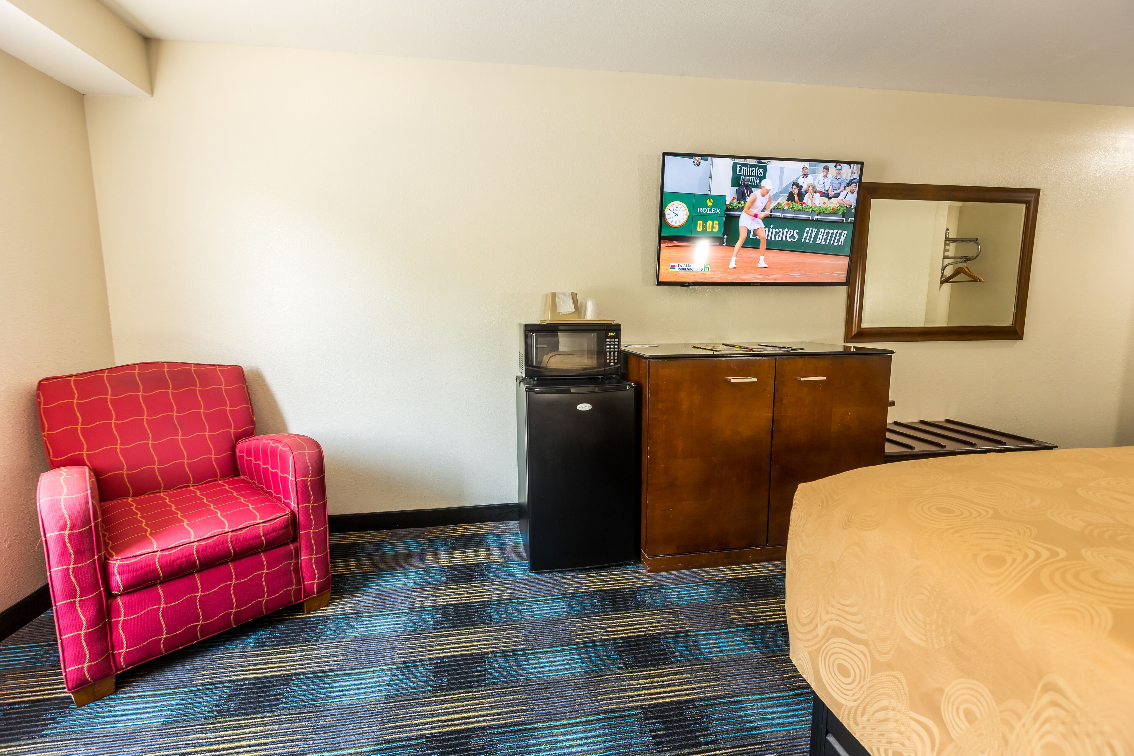 Ein Hotelzimmer mit einem roten Sessel, einem an der Wand montierten Fernseher, einer Minibar und einem Bett mit gemustertem Überwurf.