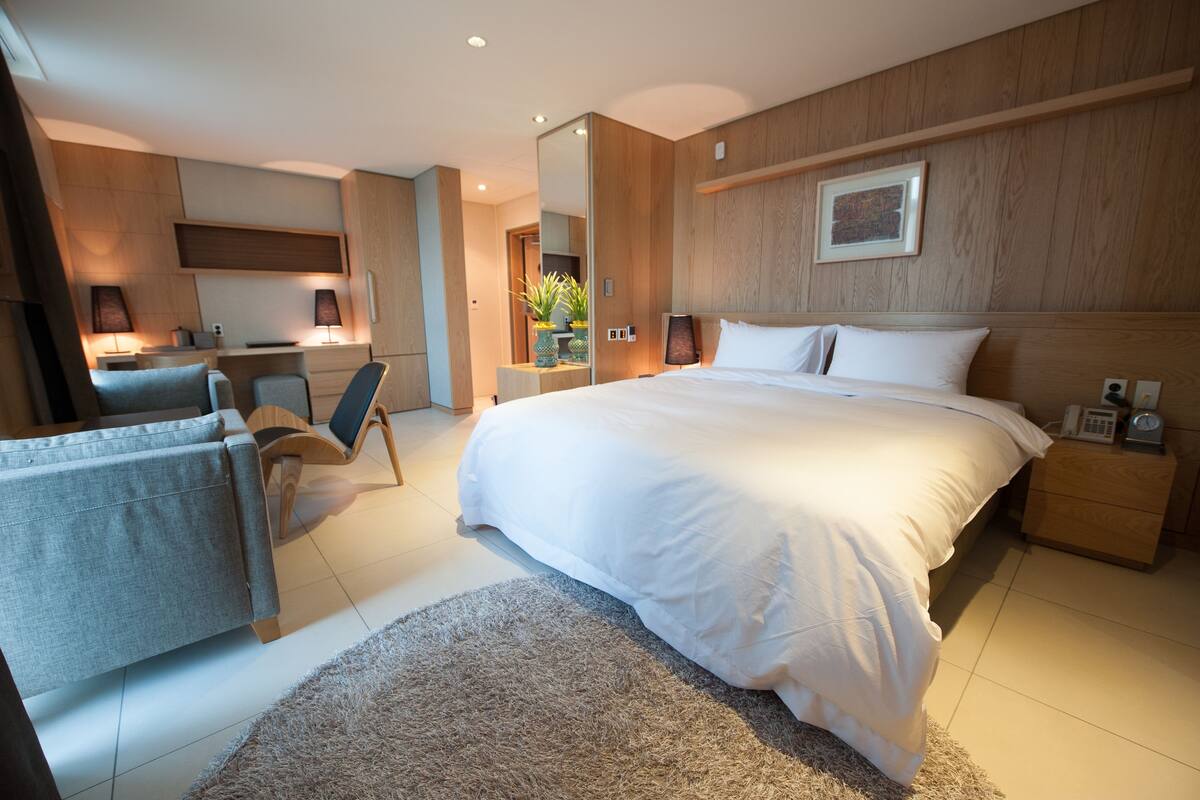 A modern hotel room with a large bed, a desk, a chair, and a small bathroom area.