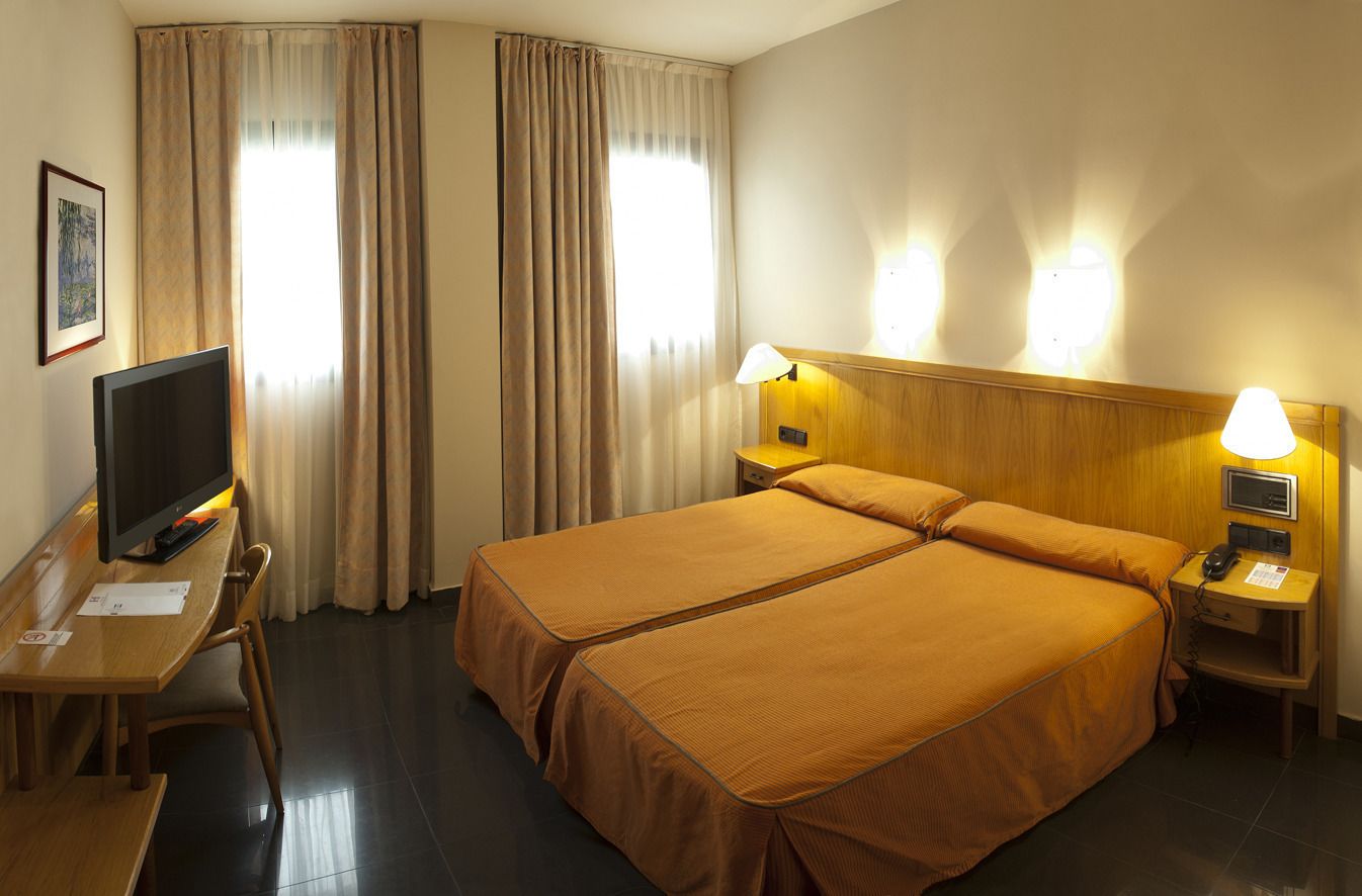 Habitación de hotel con cama doble, escritorio con televisor, lámpara y mesita de noche con teléfono.