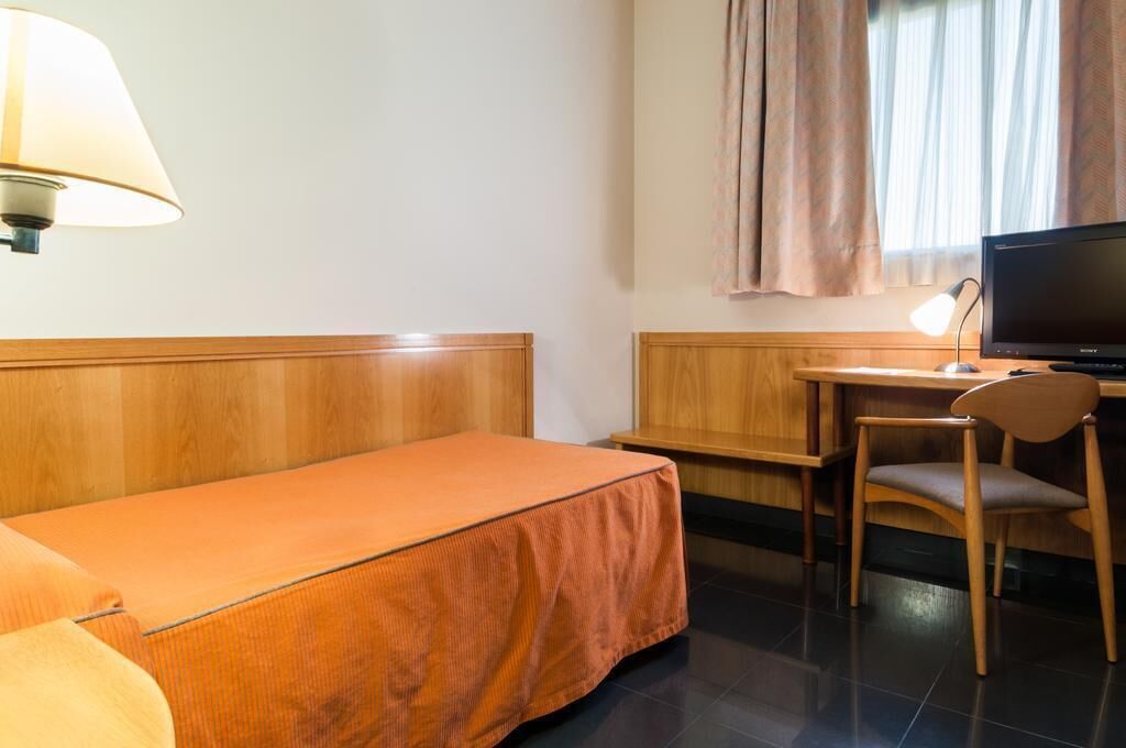 A hotel room with a single bed, a desk with a chair, a television, and a lamp.