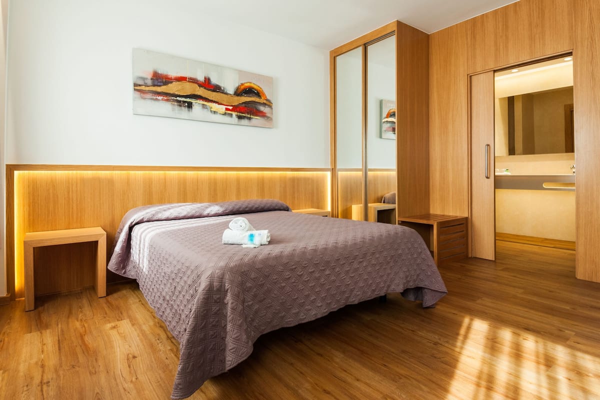 Una habitación de hotel moderna con cabecera de madera, una cama grande con una manta texturizada, una mesita de noche con una lámpara, un aparador de madera, un espejo grande y un armario de madera.