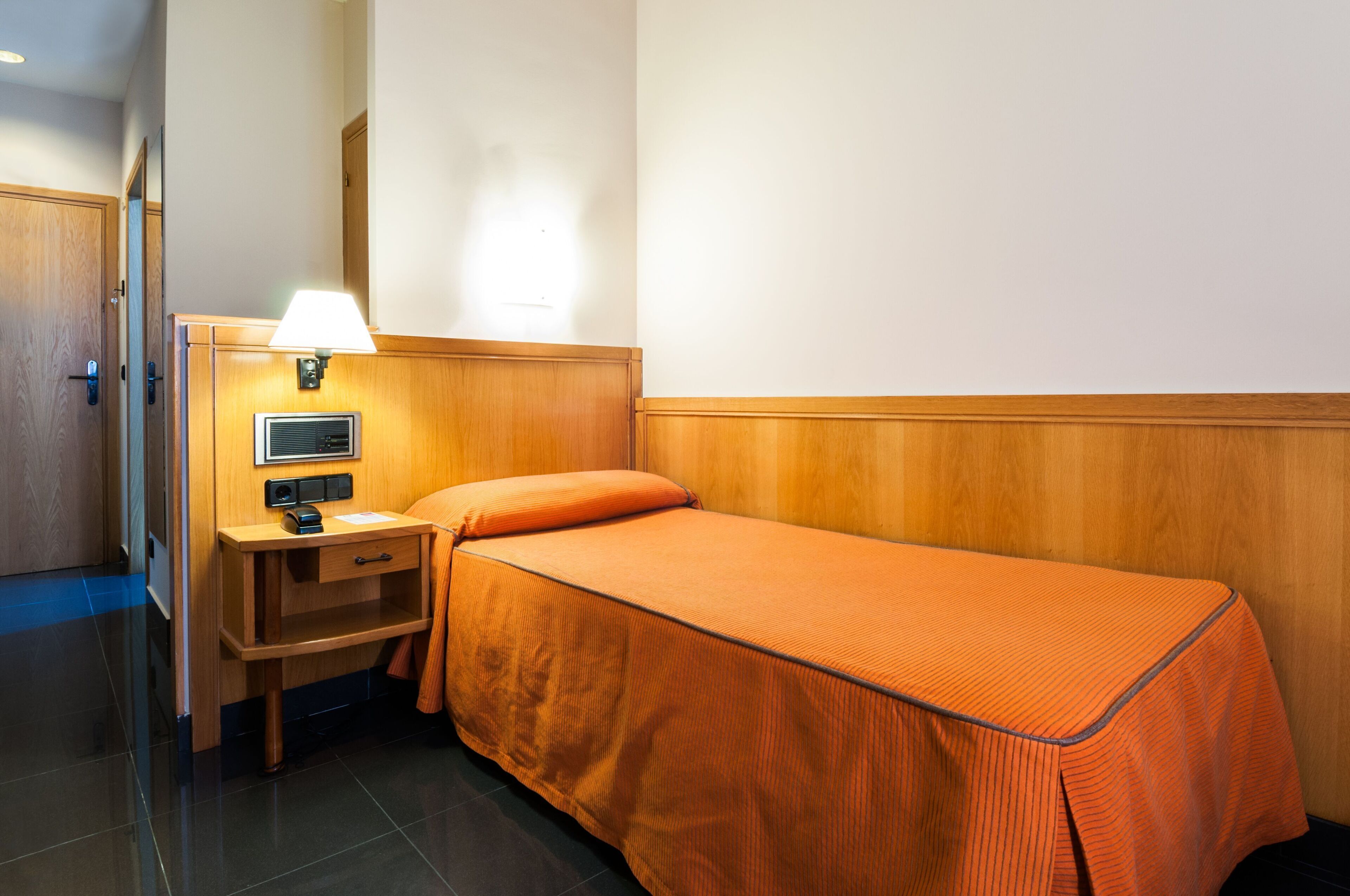 A single bed with an orange bedspread, a bedside table with a lamp, and a small drawer unit.
