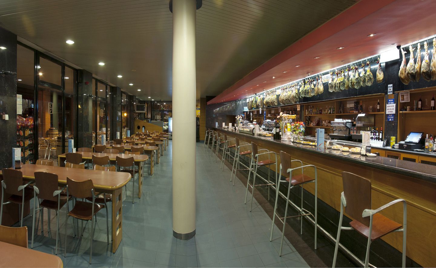 A spacious cafeteria with multiple dining tables and chairs, a central pillar, and a counter with food items and beverages.