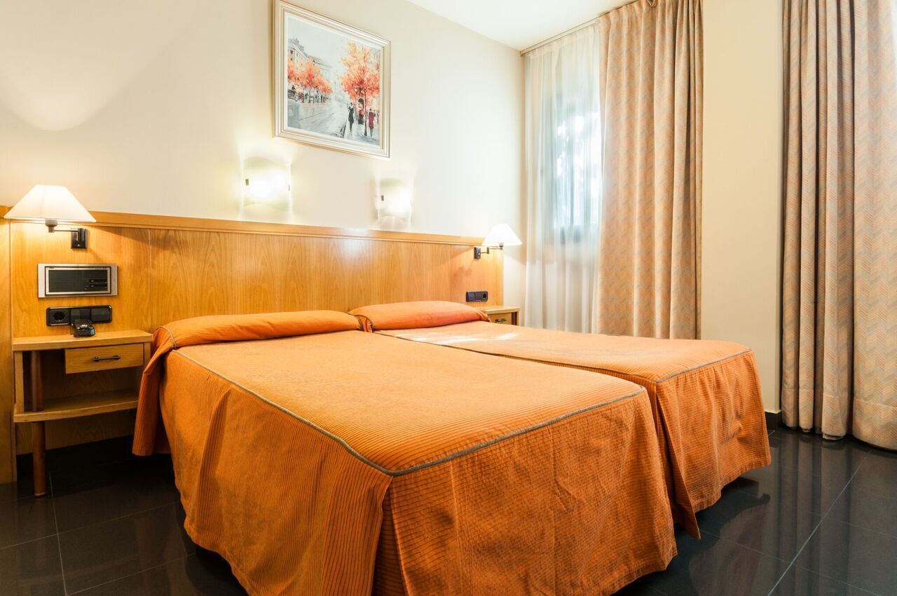 A hotel room with two beds, a wooden headboard, a nightstand with a lamp, a painting on the wall, and a window with curtains.