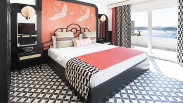 A modern hotel room with a large bed, a decorative headboard, a nightstand, and a view of the sea through large windows.