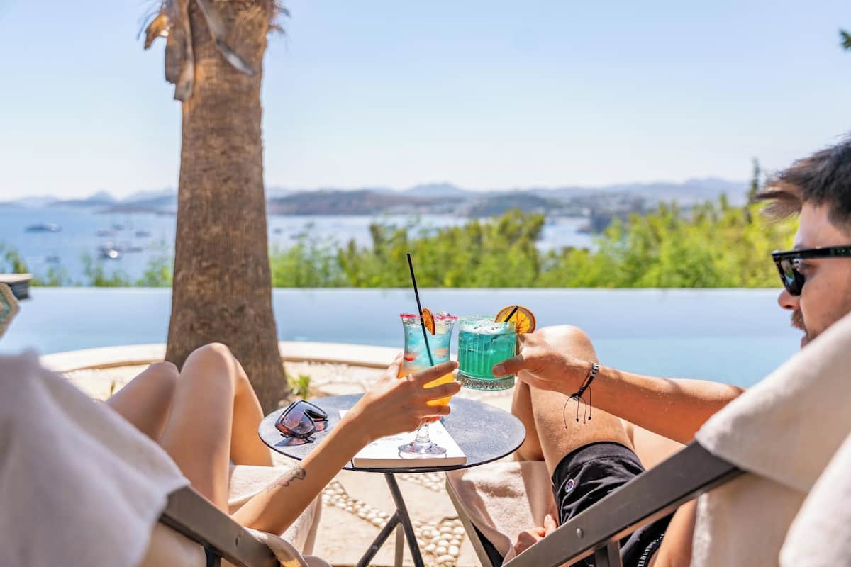 Two people sitting at a table with drinks, overlooking a body of water and mountains.