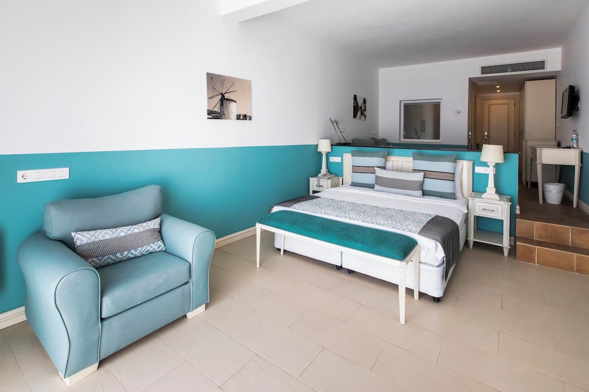 A bedroom with a bed, a chair, a nightstand, a lamp, and a wall-mounted TV.