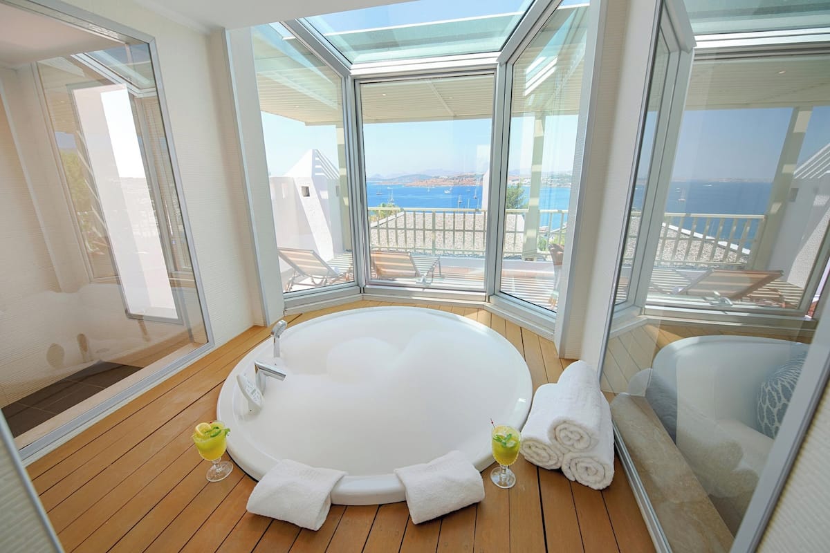 A modern bathroom with a large bathtub, wooden flooring, and a view of the sea through large windows.