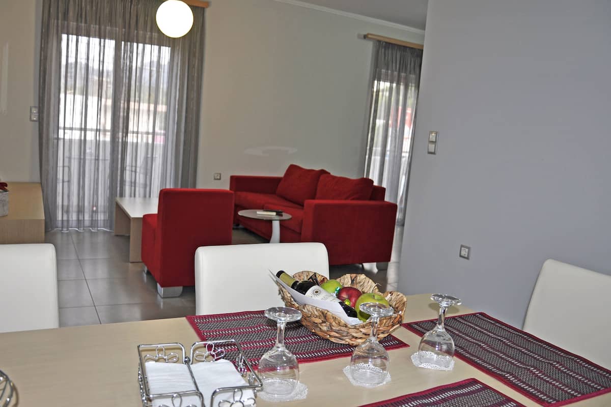 A modern living room with a red sofa, a dining table with a basket of fruit and wine glasses, and a large window with curtains.