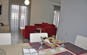 A modern living room with a red sofa, a dining table with a basket of fruit and wine glasses, and a large window with curtains. - Daniel Luxury Apartments (Rhodes)