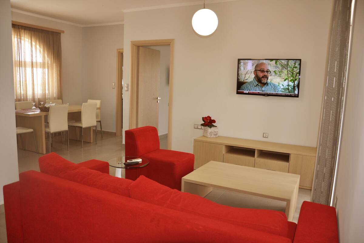 A modern living room with red sofas, a glass coffee table, a TV mounted on the wall, and a dining area with a wooden table and chairs.