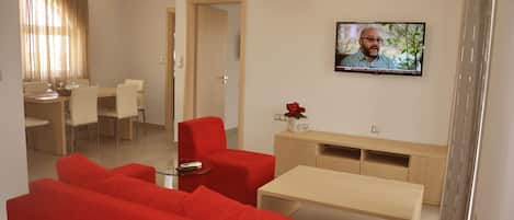 A modern living room with red sofas, a glass coffee table, a TV mounted on the wall, and a dining area with a wooden table and chairs.